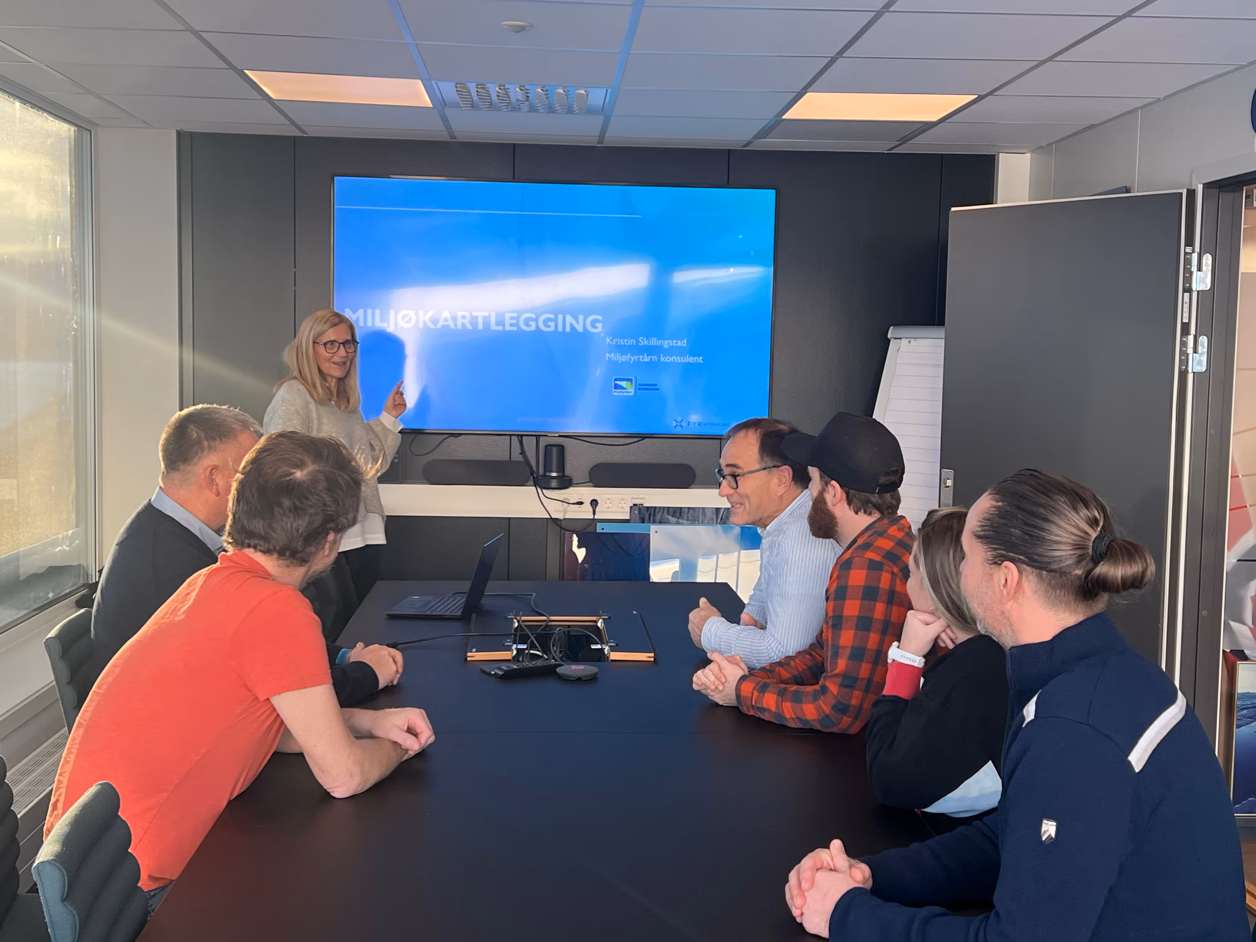 Kvinner som holder en presentasjon for fem sittende personer i et møterom med skjerm som viser tekst om miljøkartlegging.