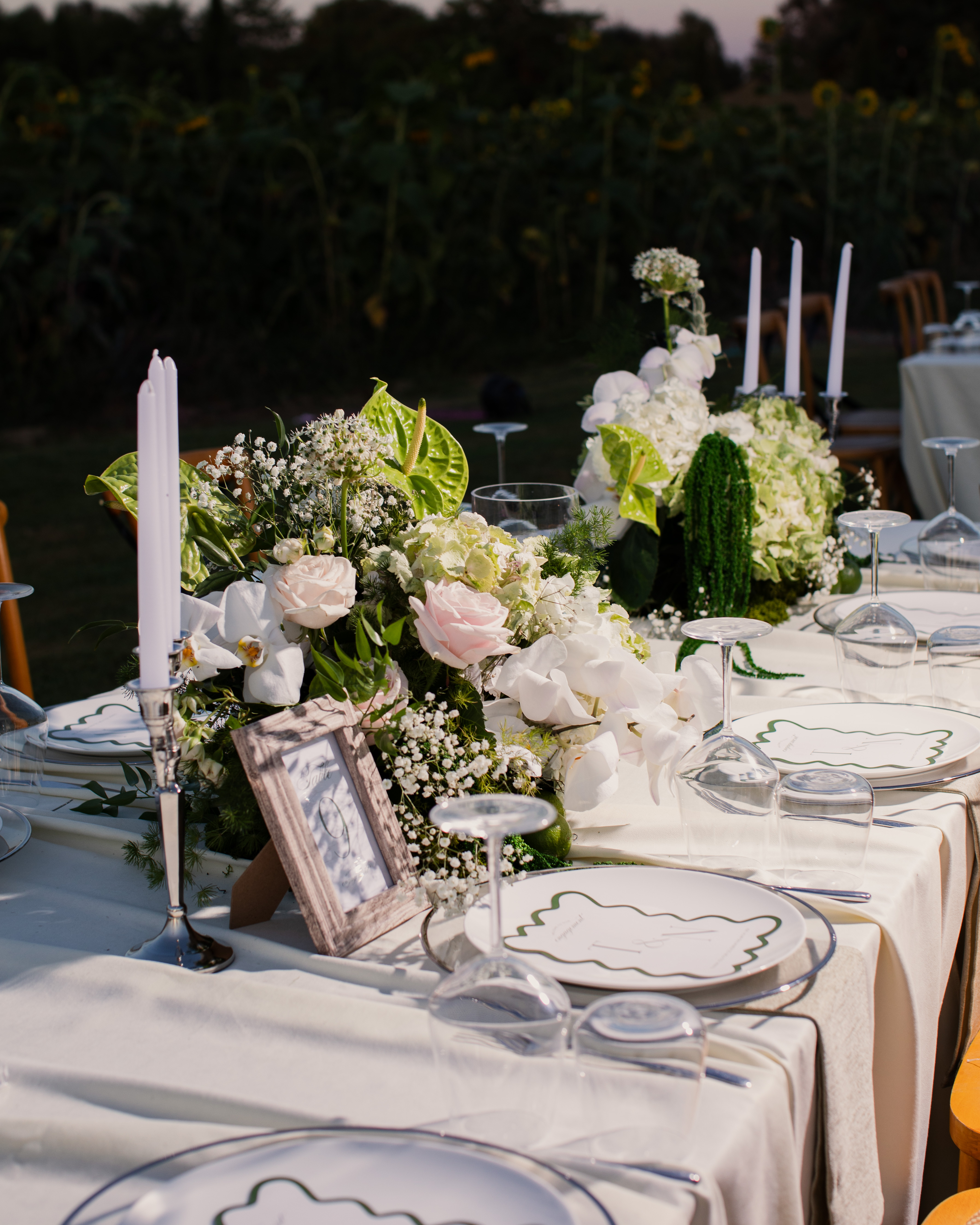 Elegant outdoor dining table with white floral centerpieces, tall white candles, and place settings with glassware and menu cards.