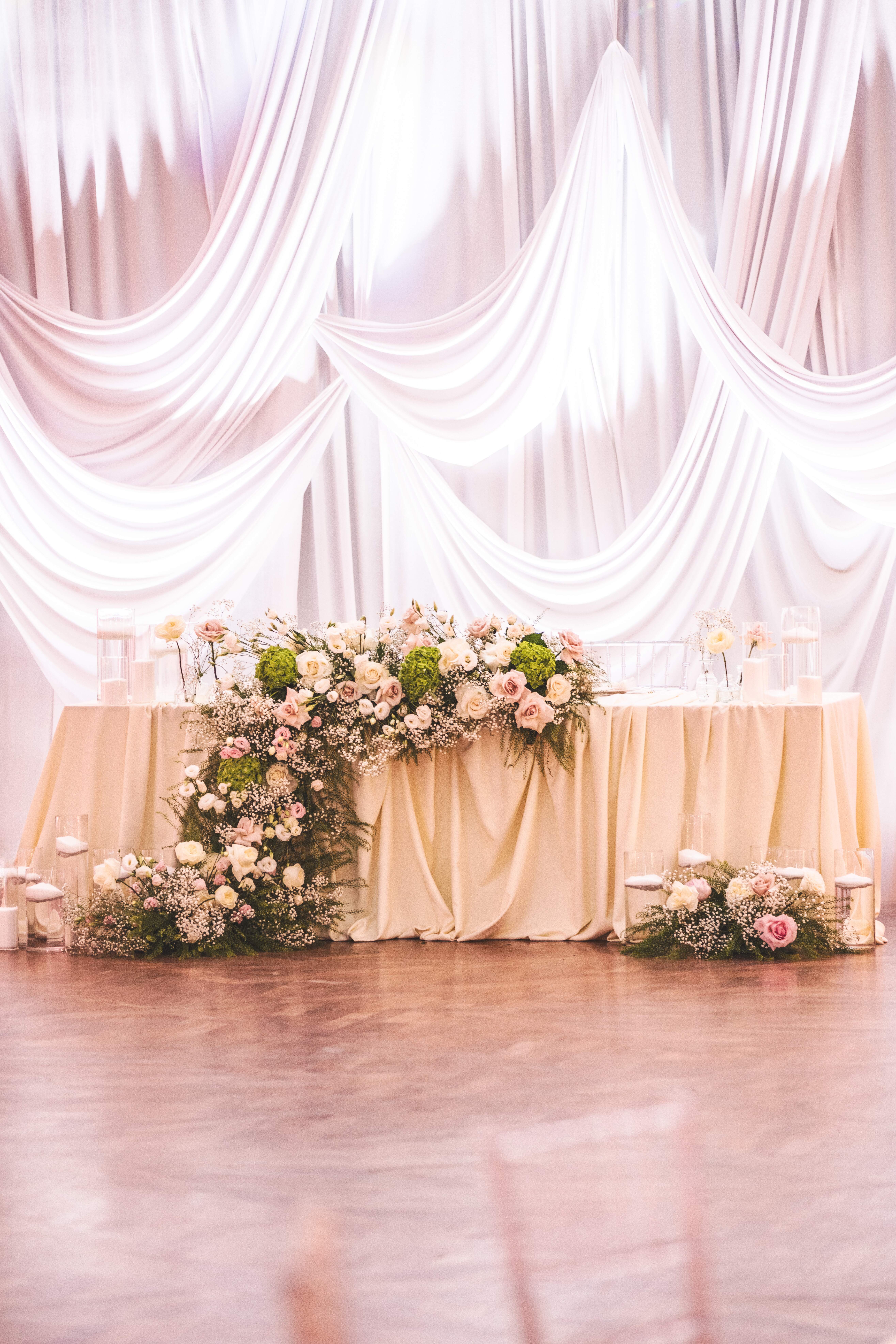 Elegant wedding head table decorated with cream-colored fabric, cascading floral arrangement of white and pink roses, greenery, and baby's breath, with glass candle holders on a wooden floor.