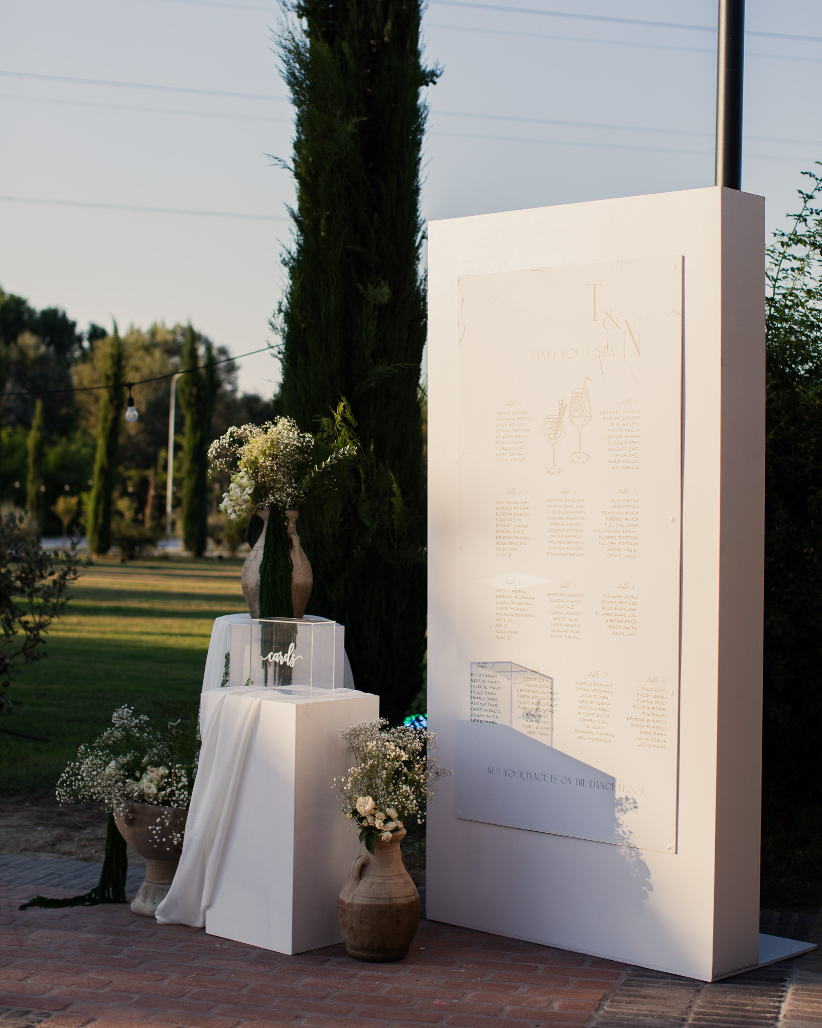 Elegant outdoor wedding seating chart display on a large white board with floral arrangements in vases and a clear box labeled 'cards' on white pedestals.