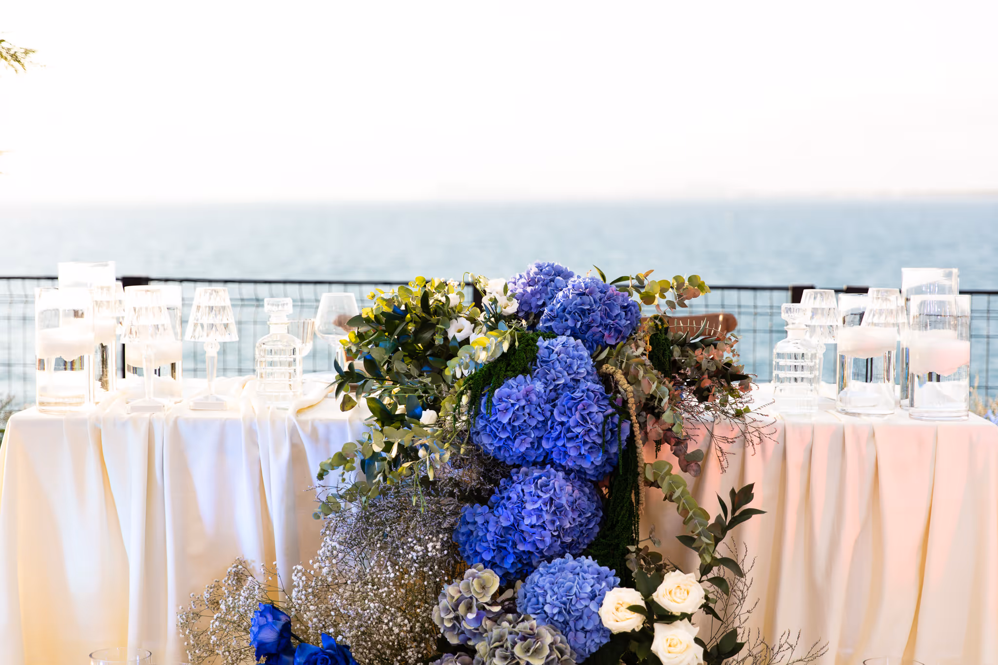 Table decorated with white cloth, blue hydrangea floral arrangement, glassware, and a sea view in the background.