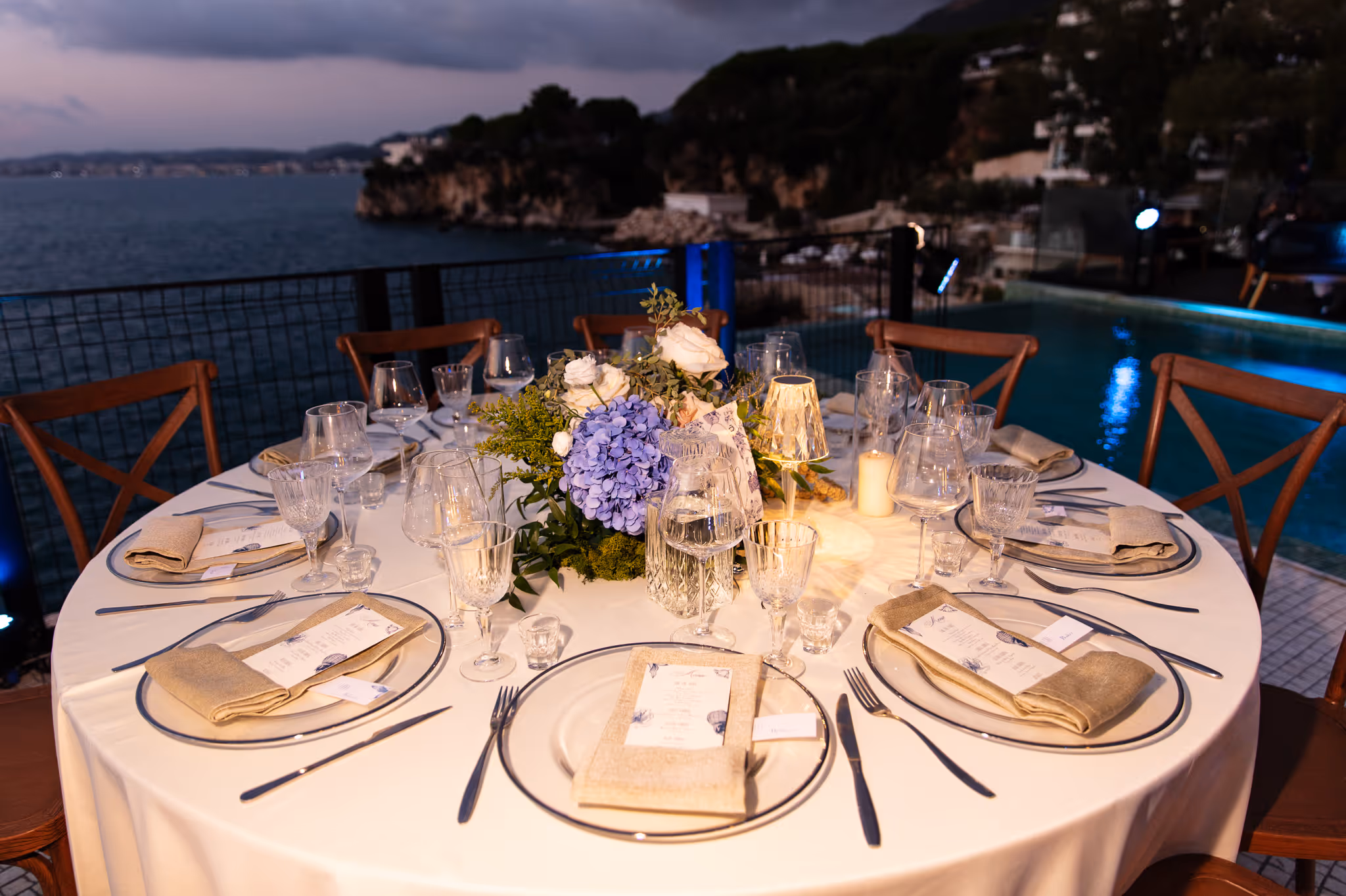 Elegant round table set for six with beige napkins, menus, glassware, and a floral centerpiece by a pool overlooking a coastal cliff at dusk.