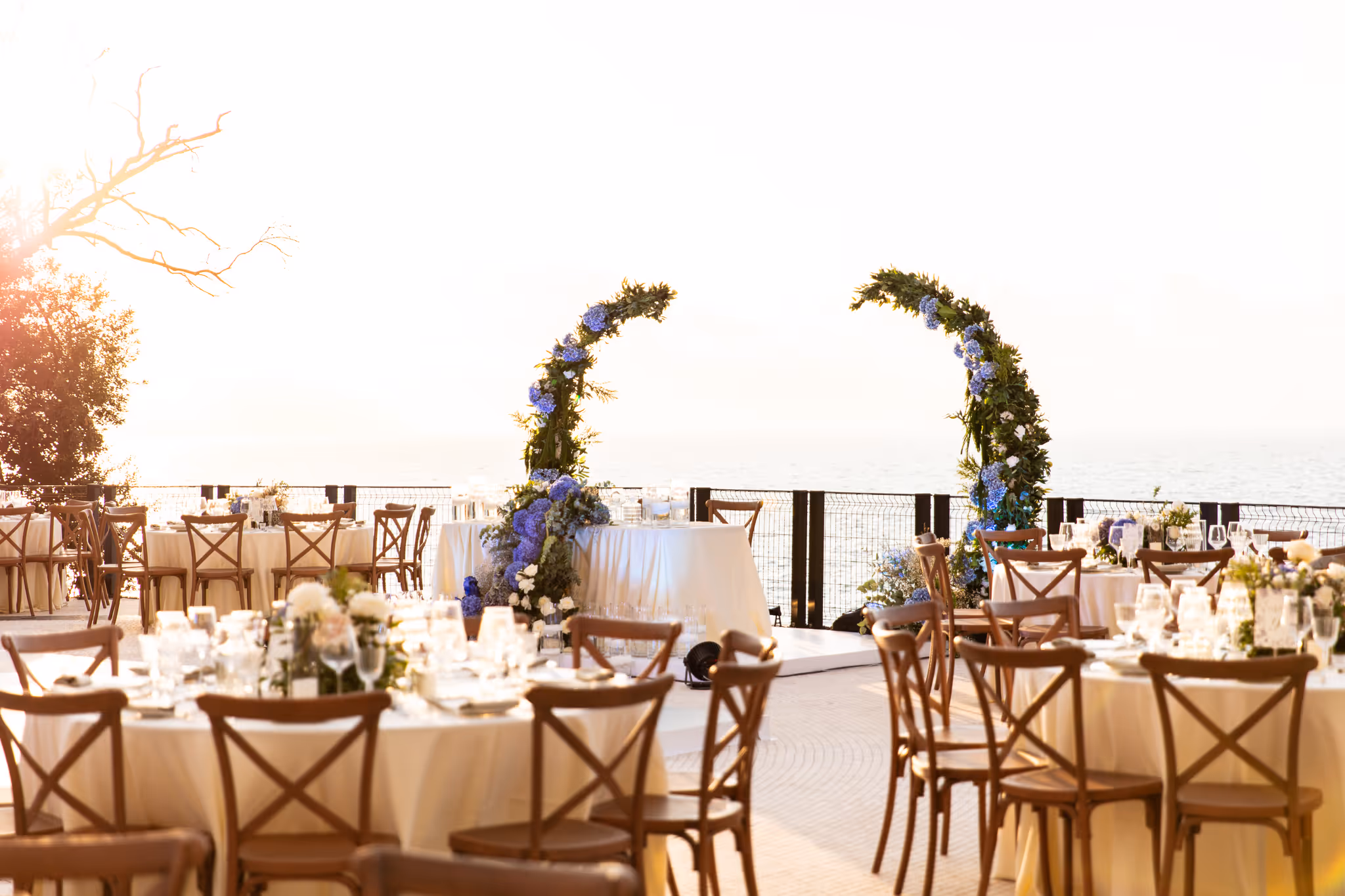 Outdoor wedding reception setup with round tables, wooden chairs, floral centerpieces, and a backdrop of the sea under soft sunlight.
