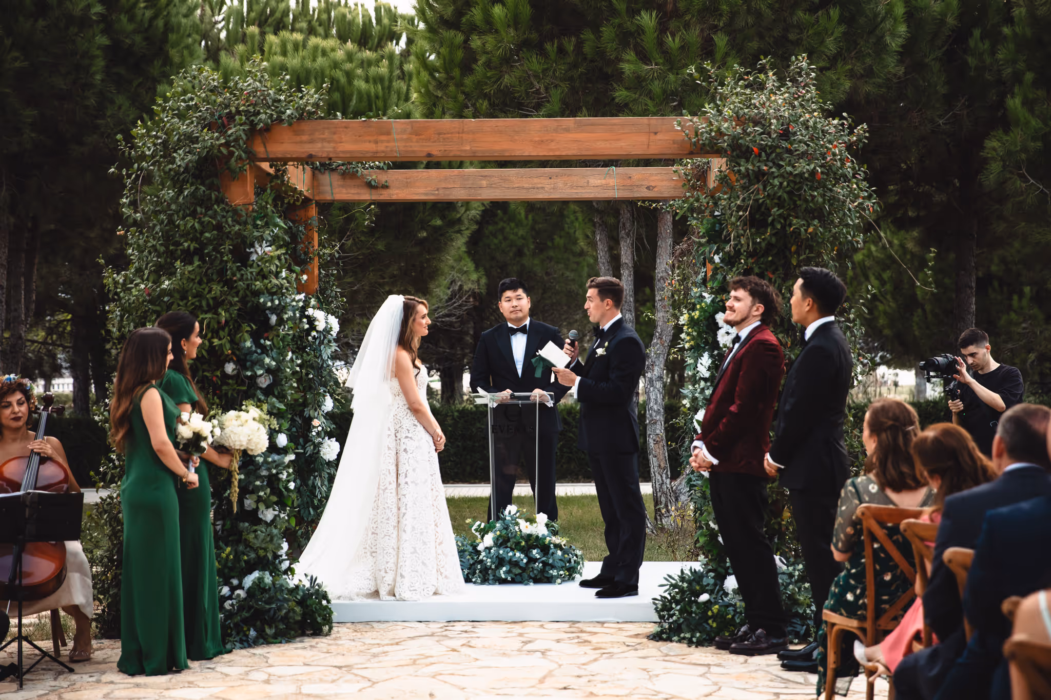Bride and groom exchanging vows under a wooden arch with greenery during an outdoor wedding ceremony surrounded by bridal party and guests.