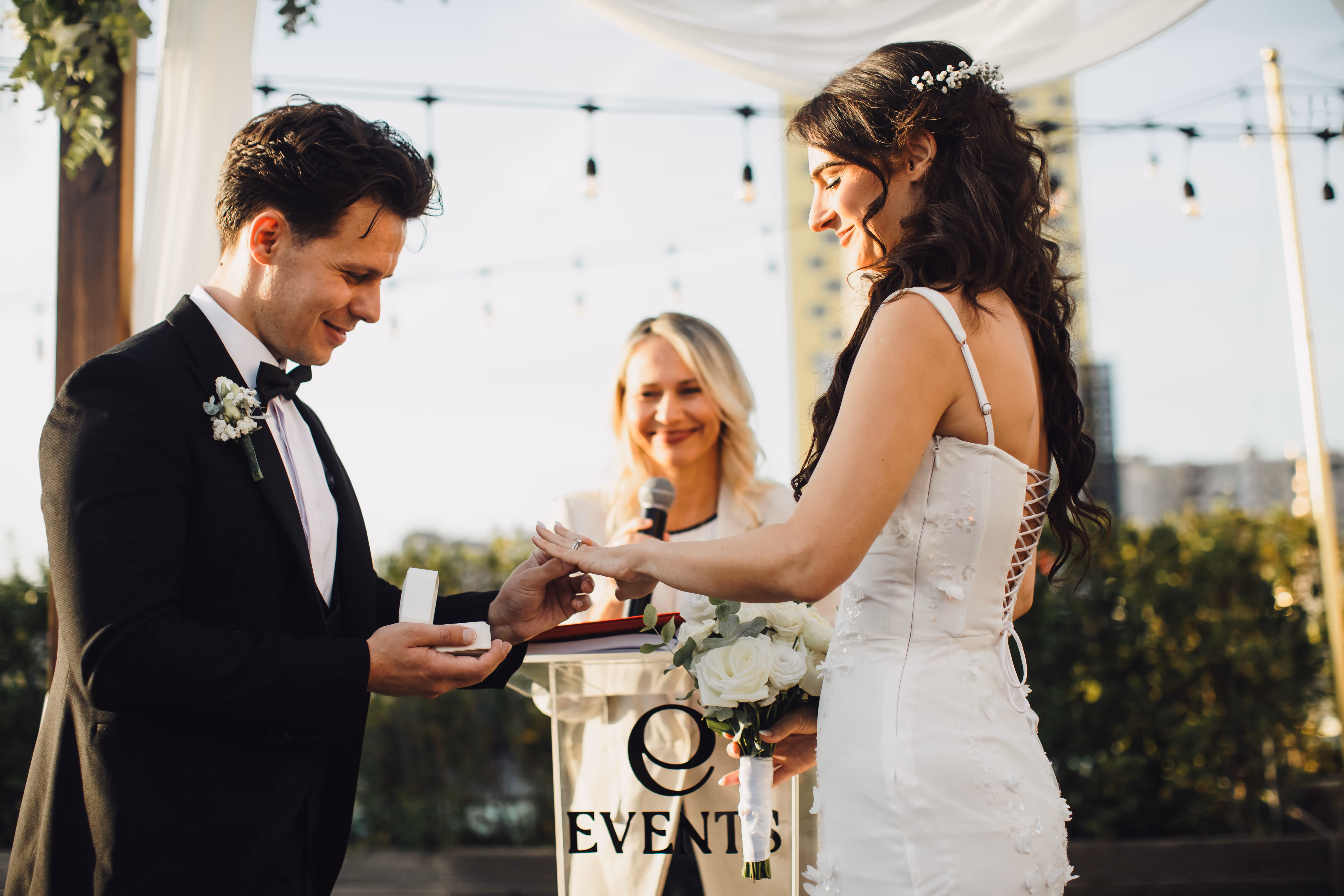 Groom placing a ring on the bride's finger during an outdoor wedding ceremony with an officiant smiling in the background.