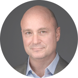 Headshot of a bald man wearing a blue shirt and gray blazer against a dark background.