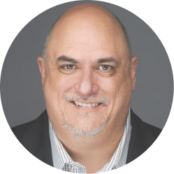 Smiling middle-aged bald man with gray goatee wearing a checkered shirt and dark blazer against a gray background.