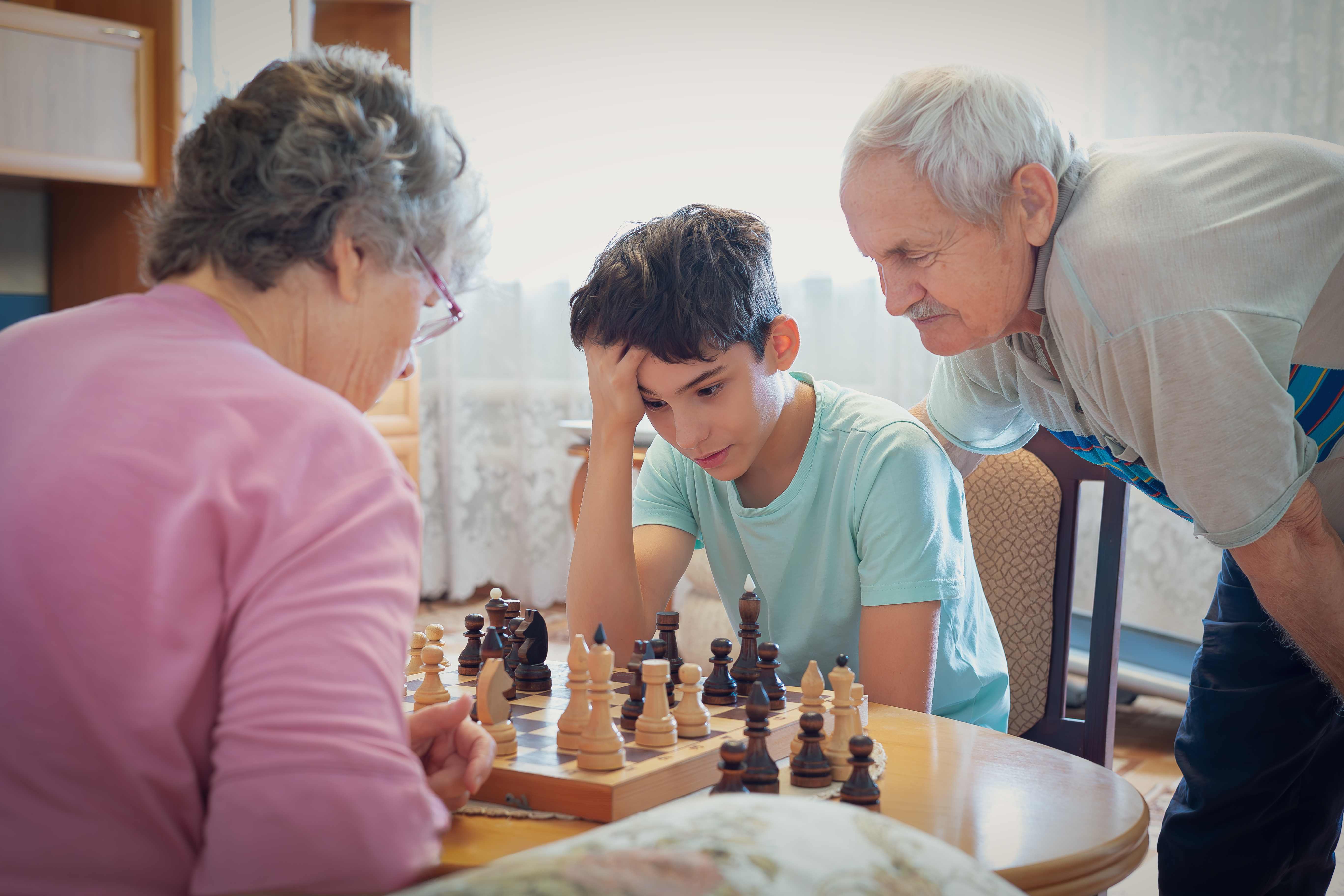 Pre-teen boy looking focused with his hand on his hair while playing chess with an elderly man and woman at a dining table