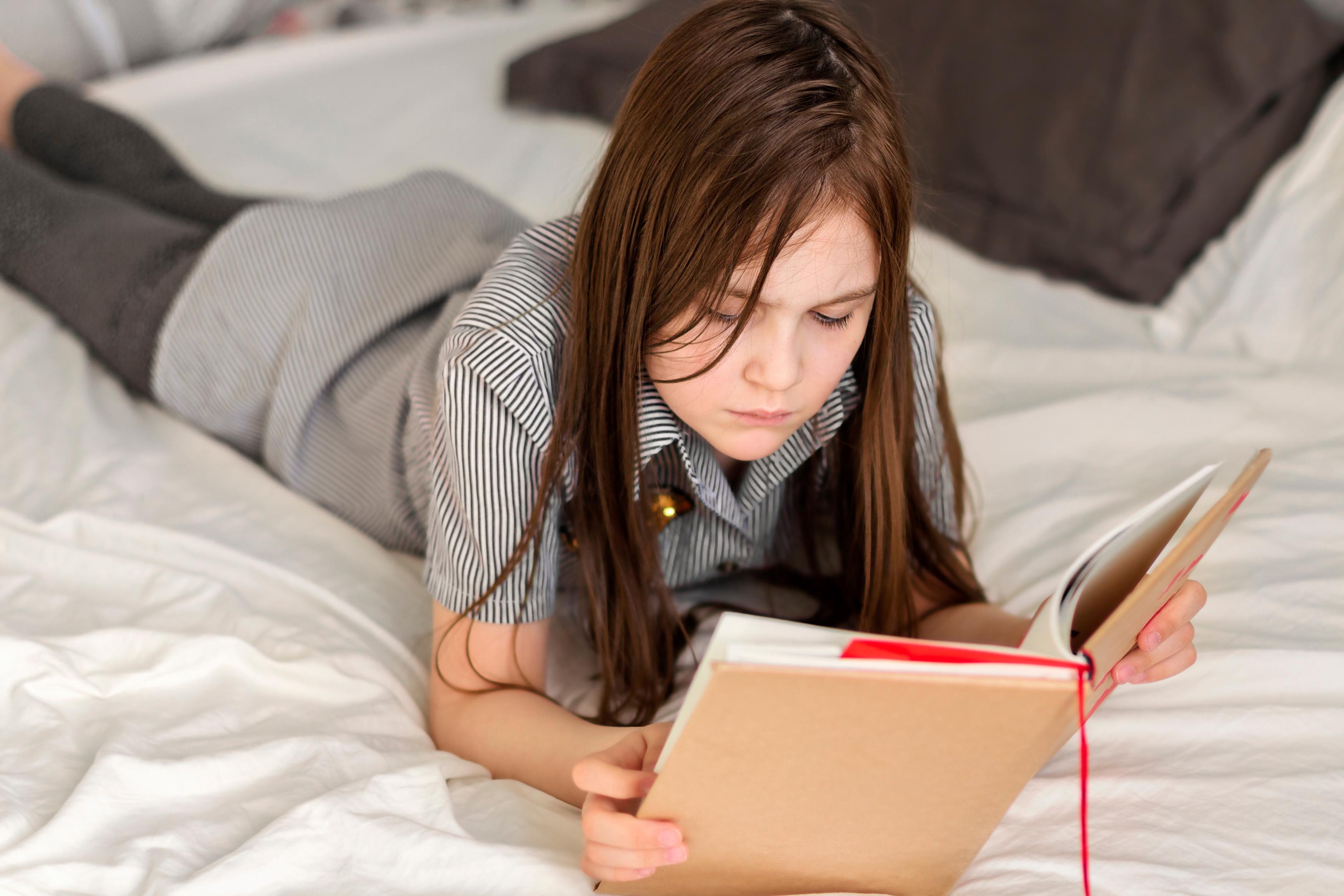 Pre-teen girl in school uniform, looking studious while lying on a bed and reading out of a hardback book