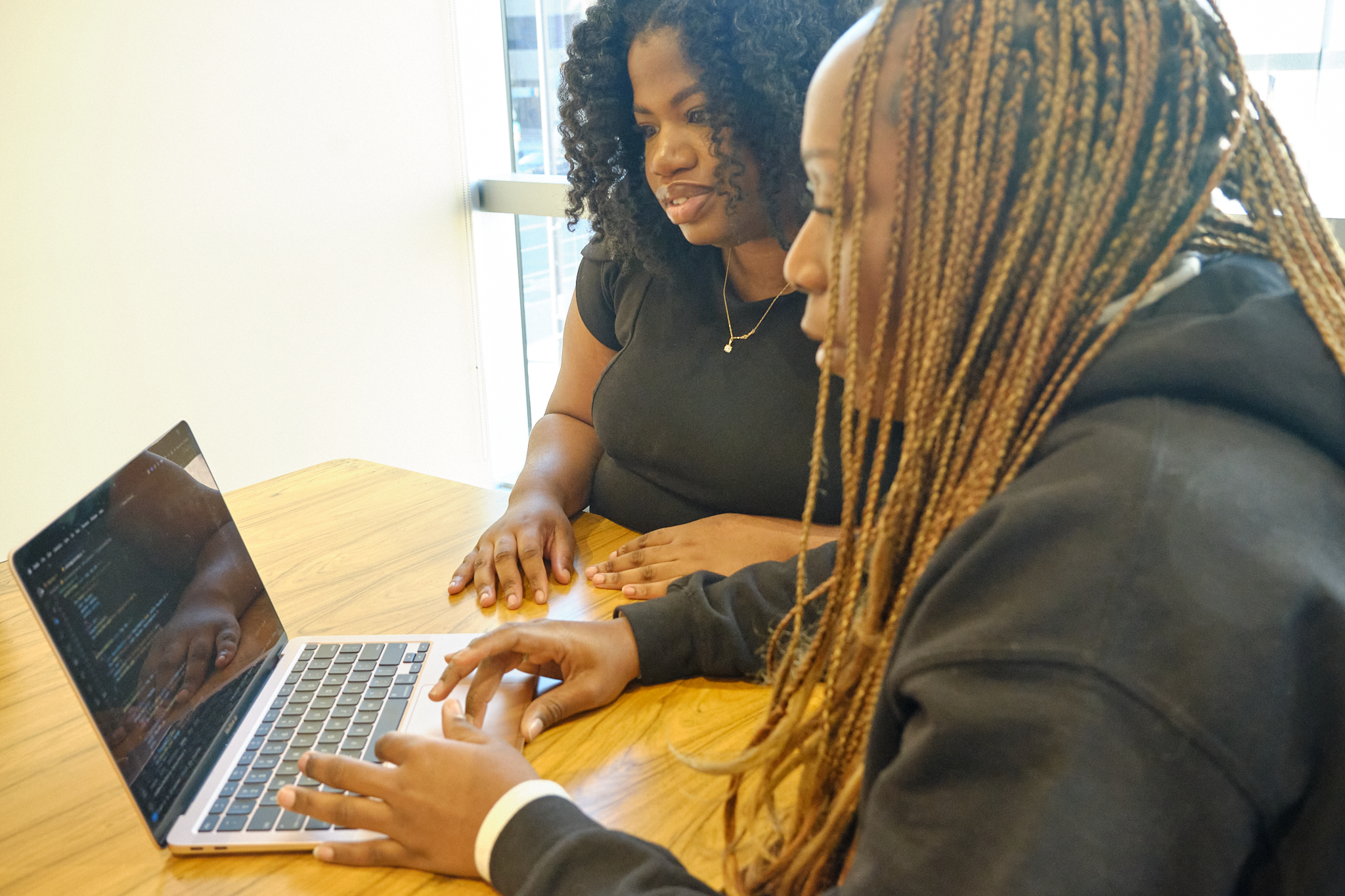 Two woman sitting, coding together in front of one computer.