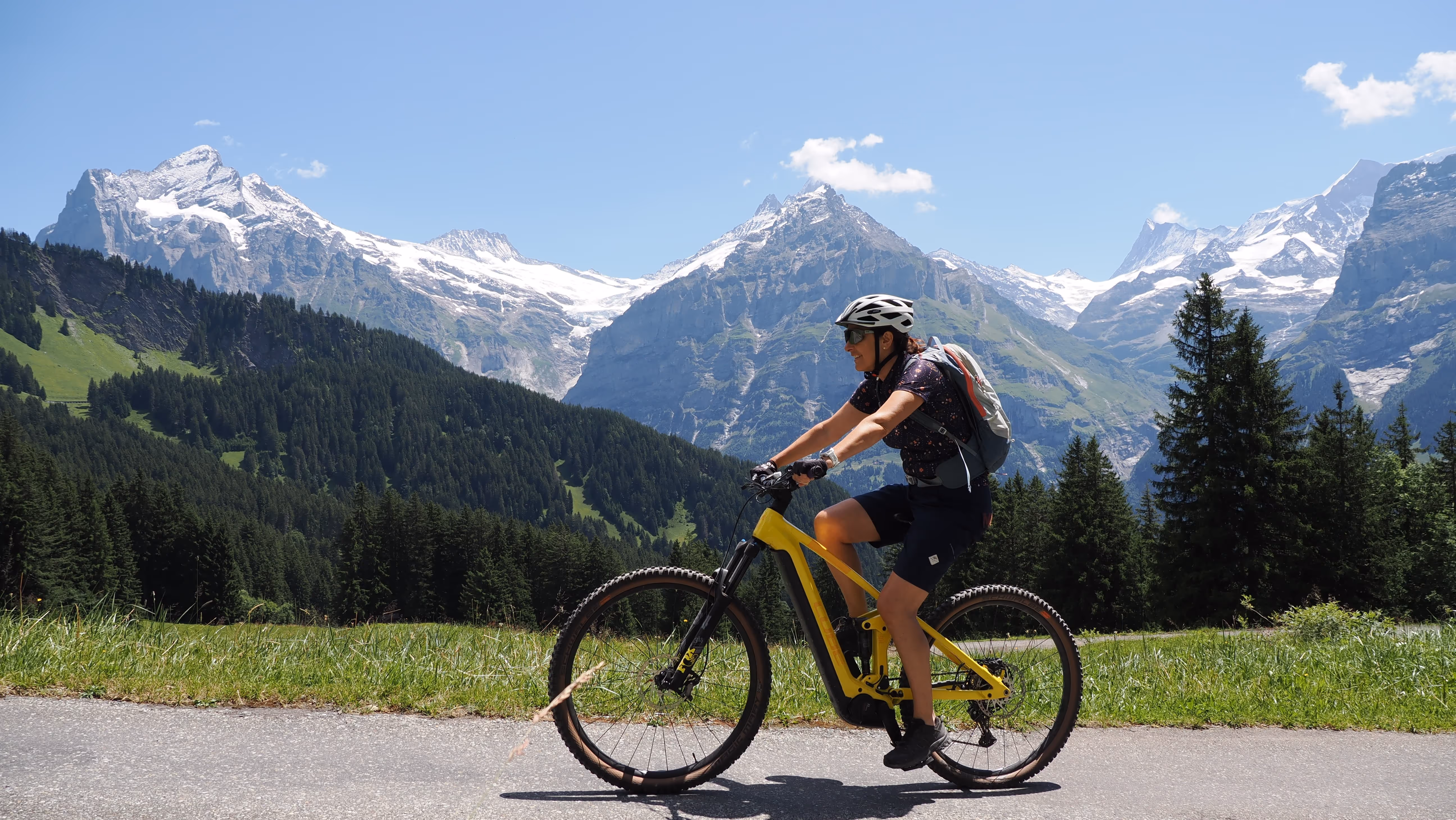 Frau fährt mit gelbem Mountainbike auf Straße vor schneebedeckten Alpenbergen und Wald unter blauem Himmel.