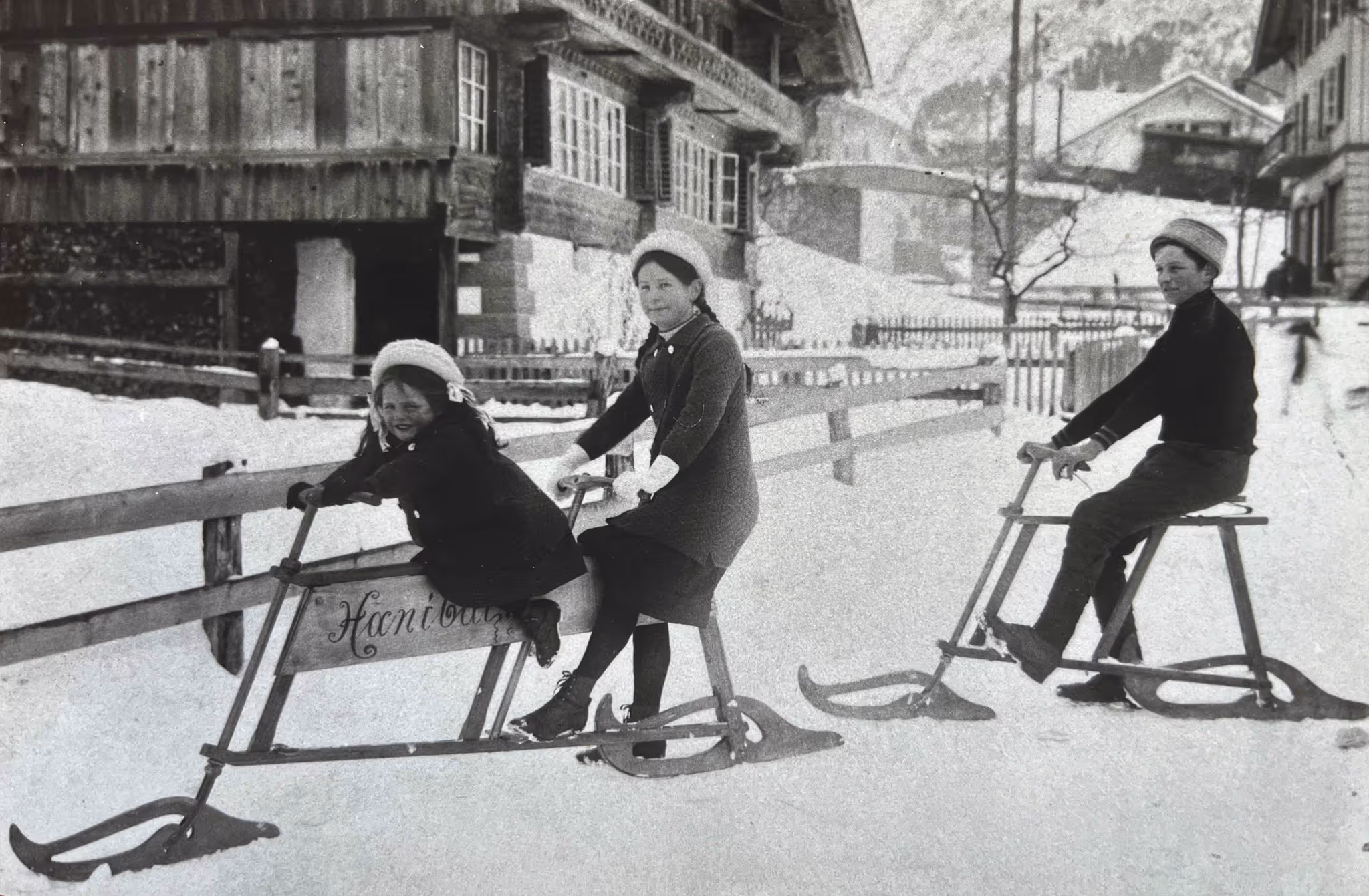 Two girls and a boy ride on traditional wooden sledges in the snow in a village with wooden houses.