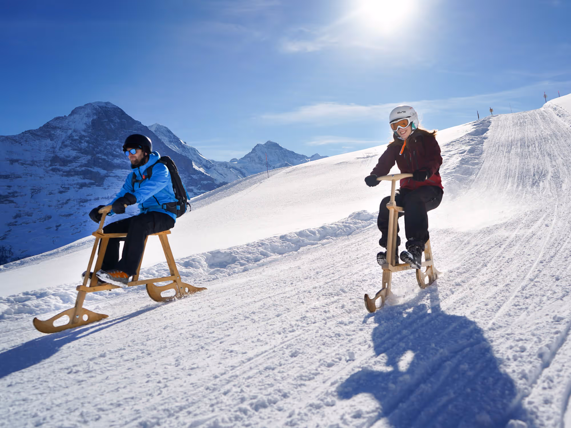 Zwei Menschen fahren auf Holzschlitten einen schneebedeckten Hang mit Bergen im Hintergrund hinunter.