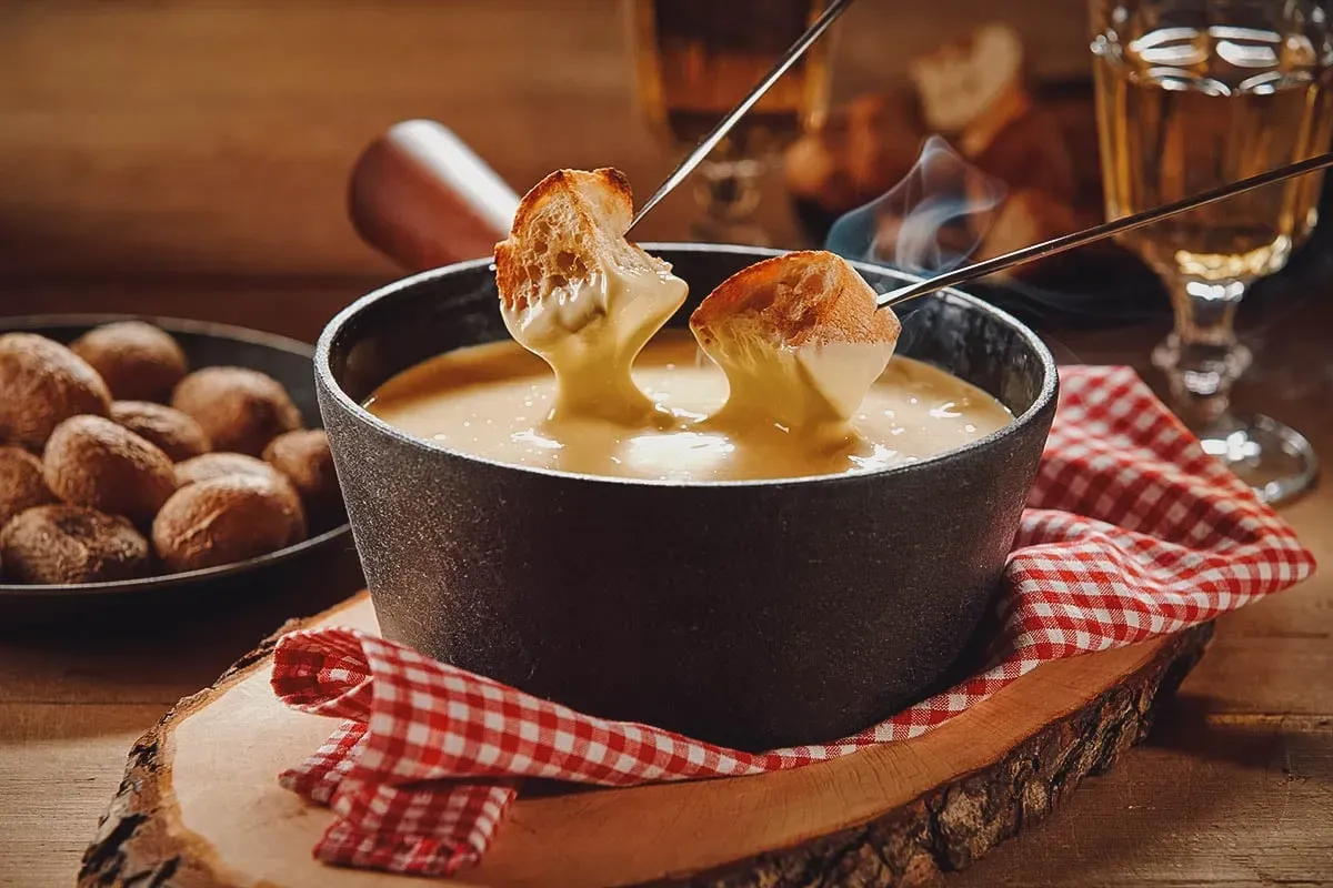 Schwarzer Topf mit geschmolzenem Käsefondue, in den zwei Gabeln mit Brot getaucht werden, auf einem Holzbrett mit rot-weiss kariertem Tuch.
