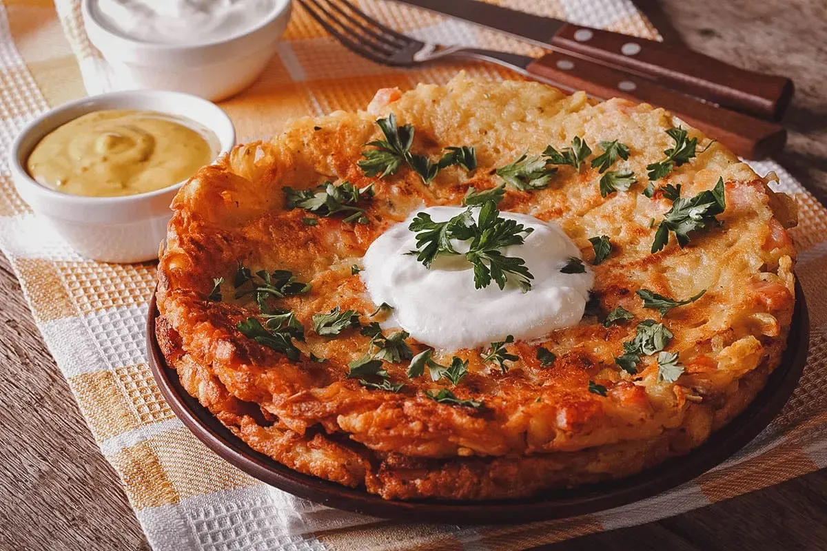Kartoffelpuffer mit saurer Sahne und frischer Petersilie auf einem braunen Teller, daneben zwei kleine Schalen mit Senf und Sauce.