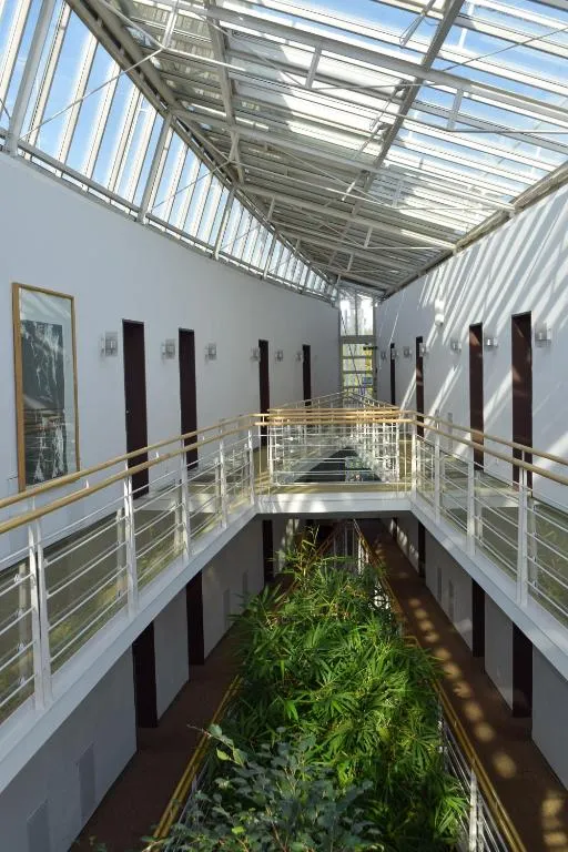 Luftige Innenansicht eines zweistöckigen Atriums, mit einem langen zentralen Pflanzbeet voller grüner Büsche im Erdgeschoss, beidseitigen Gängen mit gleichmäßig angeordneten Türen und Glas-Metall-Geländern mit Holzhandläufen, darüber ein großes schräges Glasdach, durch das Sonnenlicht Streifen und Schatten auf die weißen Wände wirft; ruhige, helle und moderne Stimmung.