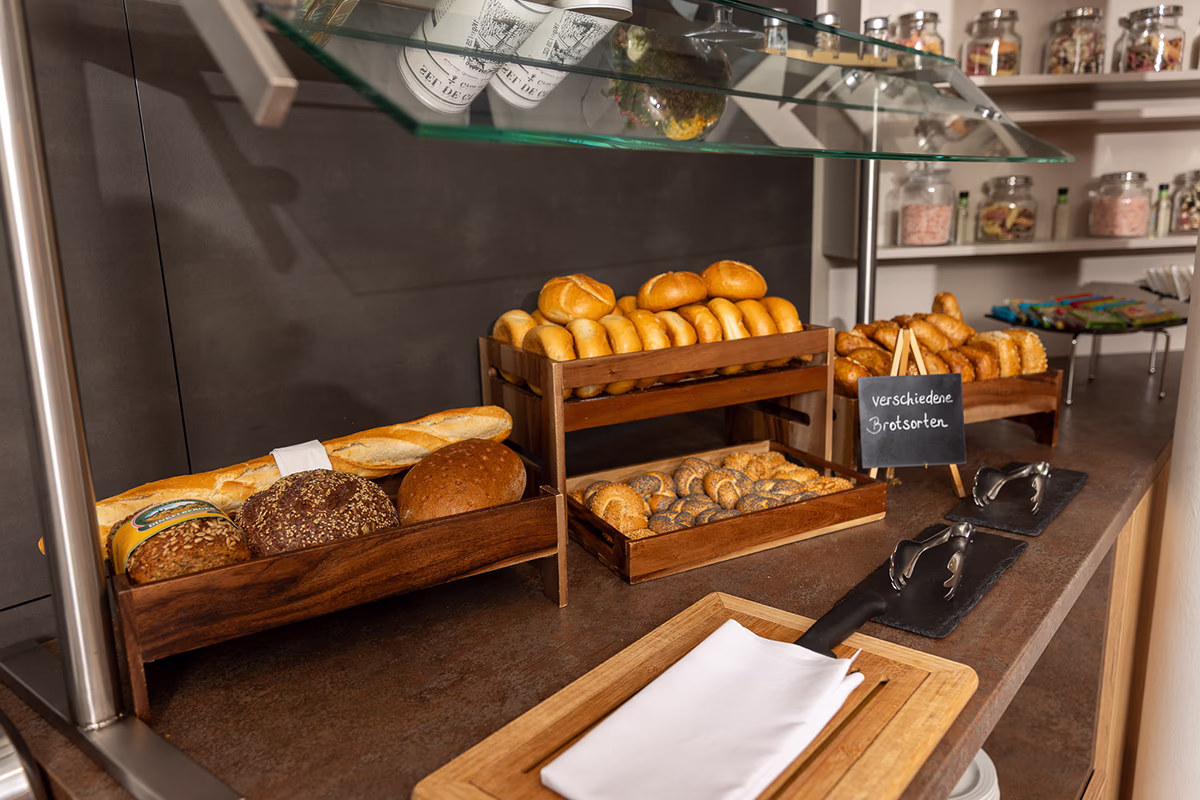 Buffetansicht im Frühstücksbereich des Scotty Hotel Hamburg: mehrere hölzerne Trays und ein zweistöckiges Holzgestell gefüllt mit Baguette, verschiedenen Körner- und Vollkornlaiben, runden Brötchen, Mohn- und Sesambrötchen sowie Croissants; vorne ein Holzbrett mit weißer Serviette, daneben zwei Metallzangen auf dunklen Schieferplatten; eine kleine Kreidetafel beschriftet mit „verschiedene Brotsorten“, darüber eine Glas-Glashaube als Spuckschutz und im Hintergrund Glasbehälter mit Cerealien. Warme Beleuchtung und ordentliche Anrichtung vermitteln eine einladende Frühstücksatmosphäre.