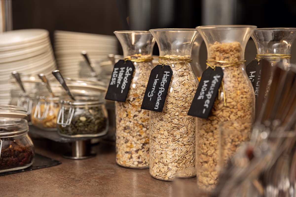 Nahaufnahme eines Frühstücksbuffets im Scotty Hotel Hamburg: drei hohe Glasflaschen mit goldfarbenem Band und schwarzen handbeschrifteten Kreidetiketten, gefüllt mit Haferflocken, Knusper‑Müsli und Schoko‑Müsli; links kleine Einmachgläser mit Trockenfrüchten und Kernen, Metalllöffel und stapelweise weiße Teller im unscharfen Hintergrund. Warme Beleuchtung und sanfte Tiefenunschärfe vermitteln eine einladende, ordentliche Frühstücksatmosphäre.