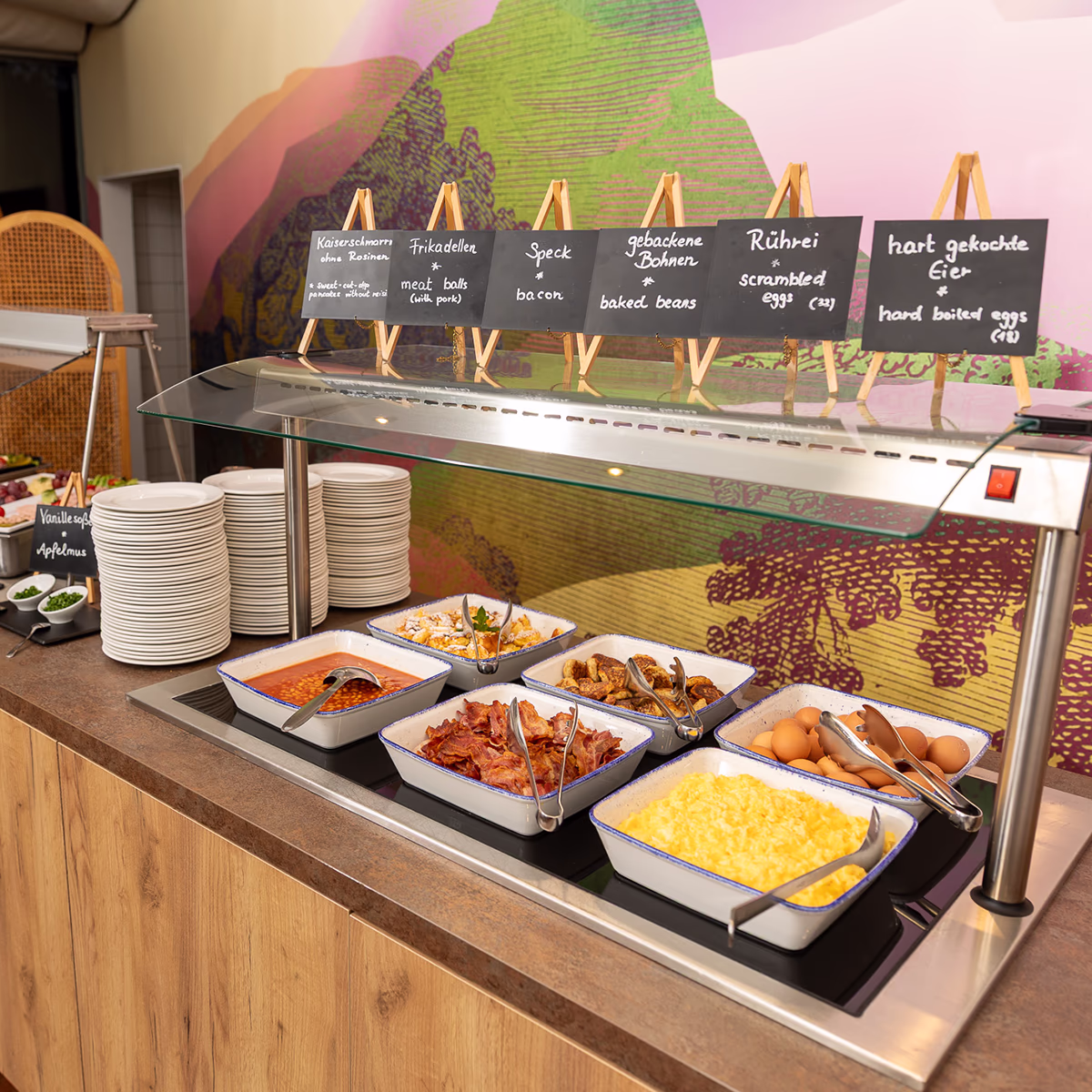 Frühstücksbuffet im Scotty Hotel Hamburg: eine lange Holztheke mit Glas-Schutz, darauf rechteckige weiße Servierschalen mit blauem Rand gefüllt mit gebackenen Bohnen, Speck, Bratkartoffeln, Rührei und hartgekochten Eiern sowie Schälchen mit Beilagen. Links gestapelte weiße Teller und kleine Kreidetafeln auf Mini-Staffeleien mit Beschriftungen über den Speisen, im Hintergrund eine farbige Wanddekoration. Ordentliche, einladende Präsentation in warmer Beleuchtung.