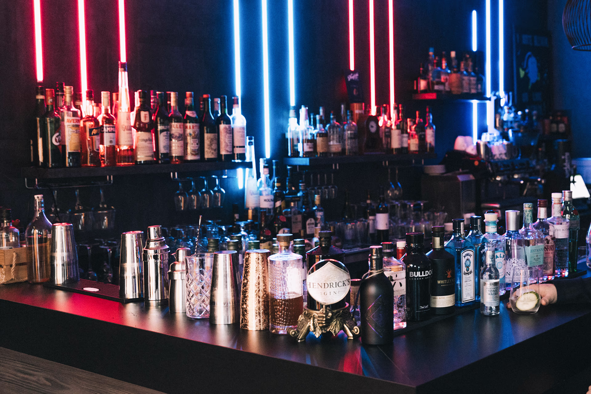 Innenaufnahme der Queen Mum Bar im Scotty Hotel Hamburg: dunkle Bartheke mit dicht nebeneinander stehenden Spirituosenflaschen und glänzenden Cocktail-Shakern im Vordergrund, eine auffällige Hendrick’s-Gin-Flasche auf einem kleinen dekorativen Ständer und ein Glas mit Zitronenscheibe am rechten Bildrand; im Hintergrund Regale mit weiteren Flaschen, vertikale rote und blaue Neonröhren tauchen die Szene in kühles, lebhaftes Licht und erzeugen ein modernes, stimmungsvolles Nachtbar-Ambiente.