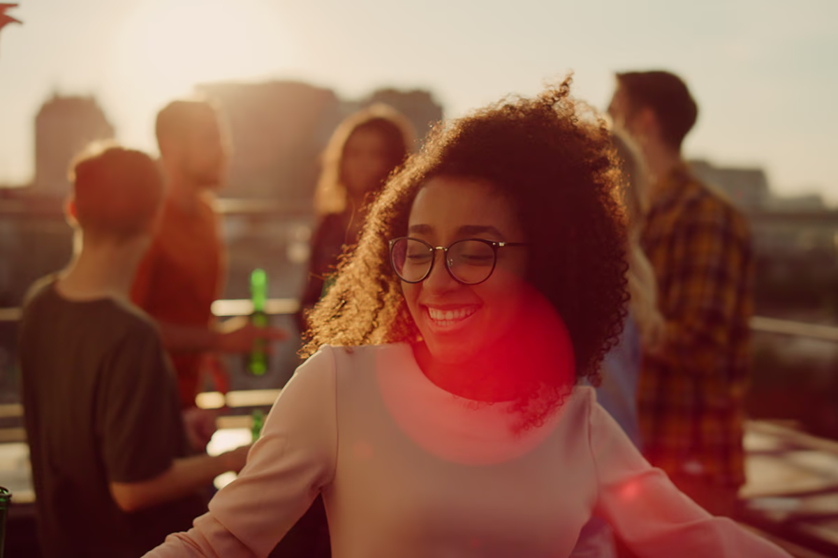 Junge Frau mit lockigem Haar und Brille steht im Vordergrund auf einer Dachterrasse bei Sonnenuntergang, lächelt mit geschlossenen Augen und bewegt sich leicht, ein roter Lichtreflex (Lensflare) zieht über Brust und Gesicht; im unscharfen Hintergrund unterhalten sich mehrere Freundinnen und Freunde und halten Flaschen, warme, fröhliche Abendstimmung.