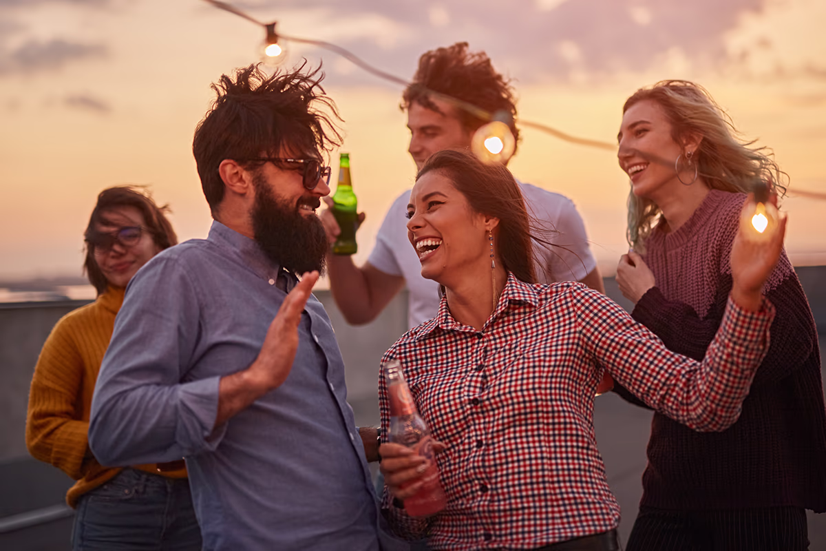 Gruppe junger Erwachsener feiert auf einer Dachterrasse bei Sonnenuntergang; im Vordergrund ein bärtiger Mann mit Sonnenbrille und eine Frau im karierten Hemd, die lachend eine Flasche hält, im Hintergrund unterhalten sich drei weitere Personen; Lichterkette und warme Abendstimmung vermitteln eine fröhliche, ausgelassene Atmosphäre.