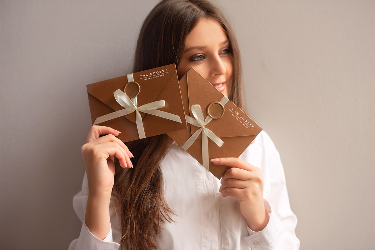 Junge Frau in weißer Bluse hält lächelnd zwei braune Geschenkumschläge mit cremefarbenen Schleifen und goldenen Siegelringen, auf denen „THE SCOTTY HOTEL HAMBURG“ steht; sie blickt zur Seite vor neutralem Hintergrund, warme und einladende Stimmung.