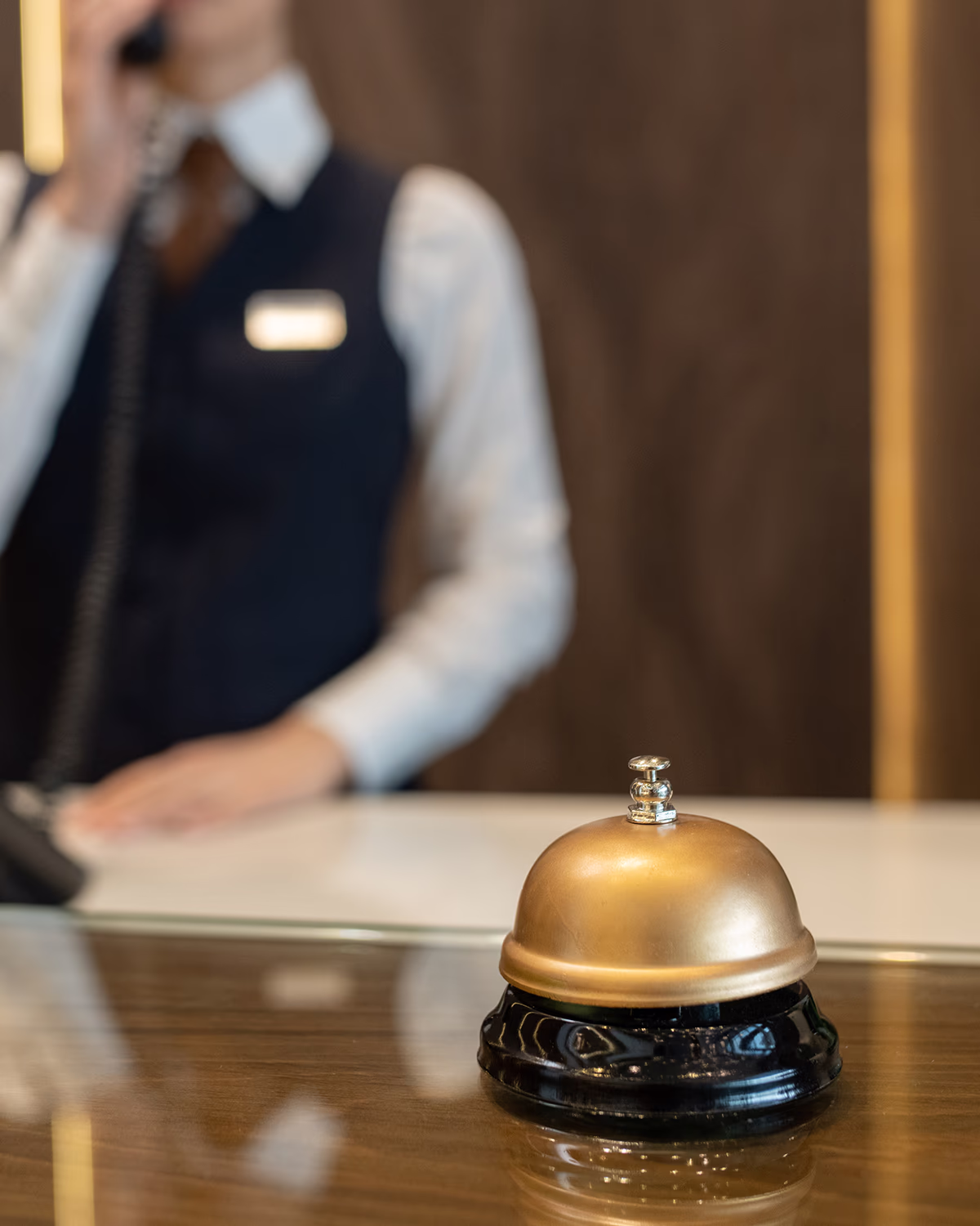 Nahaufnahme einer goldenen Empfangsglocke auf einem polierten Holztresen. Im unscharfen Hintergrund steht eine Empfangsperson in weißem Hemd und dunkler Weste und telefoniert. Warme Beleuchtung und klare, professionelle, einladende Stimmung.