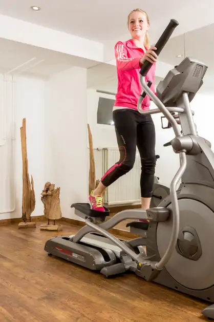 Frau in pinker Sportjacke trainiert auf einem Crosstrainer in einem hellen Fitnessraum mit Holzboden.