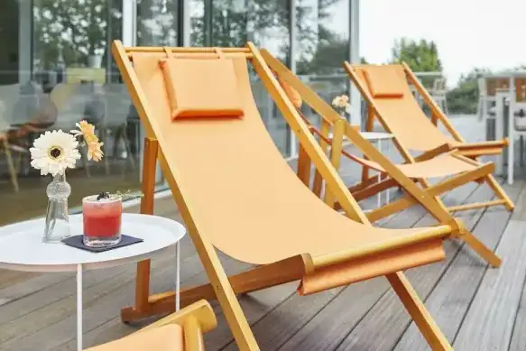 Drei orange Liegestühle aus Holz auf einer Terrasse mit zwei kleinen weißen Tischen, auf einem der Tische steht eine Vase mit Blumen und ein rotes Getränk.