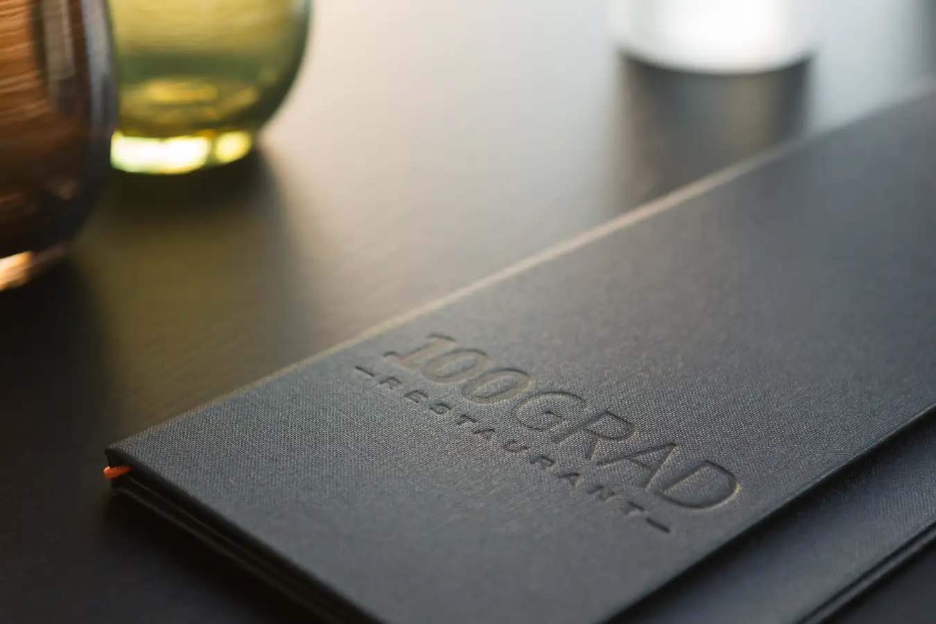 Dunkelgraue Speisekarte mit geprägtem Schriftzug 100GRAD + RESTAURANT liegt auf einem Tisch neben einem braunen, grünen und weißen Wasserglas.