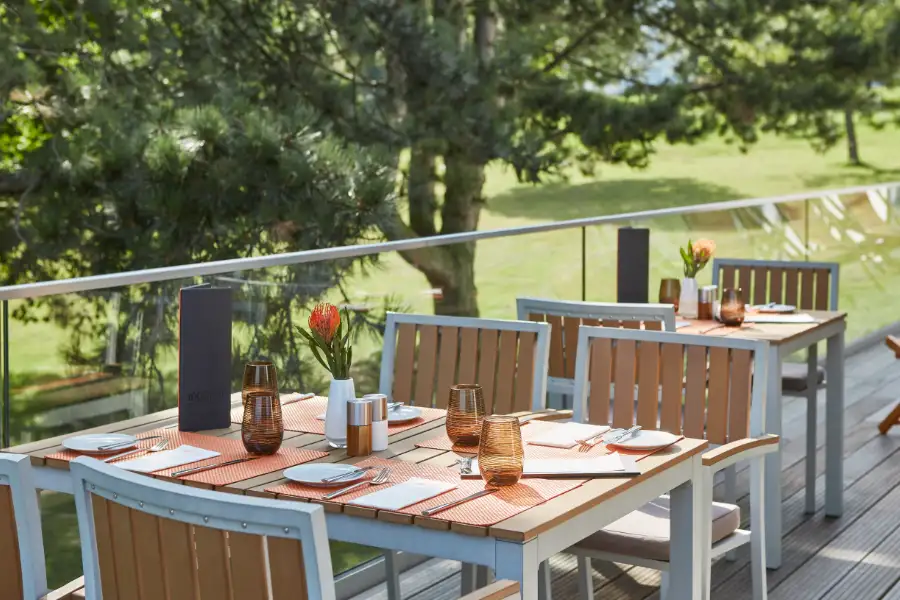 Gedeckte Tische mit orangefarbenen Platzsets und braunen Gläsern auf einer Terrasse mit Blick auf einen grünen Garten.