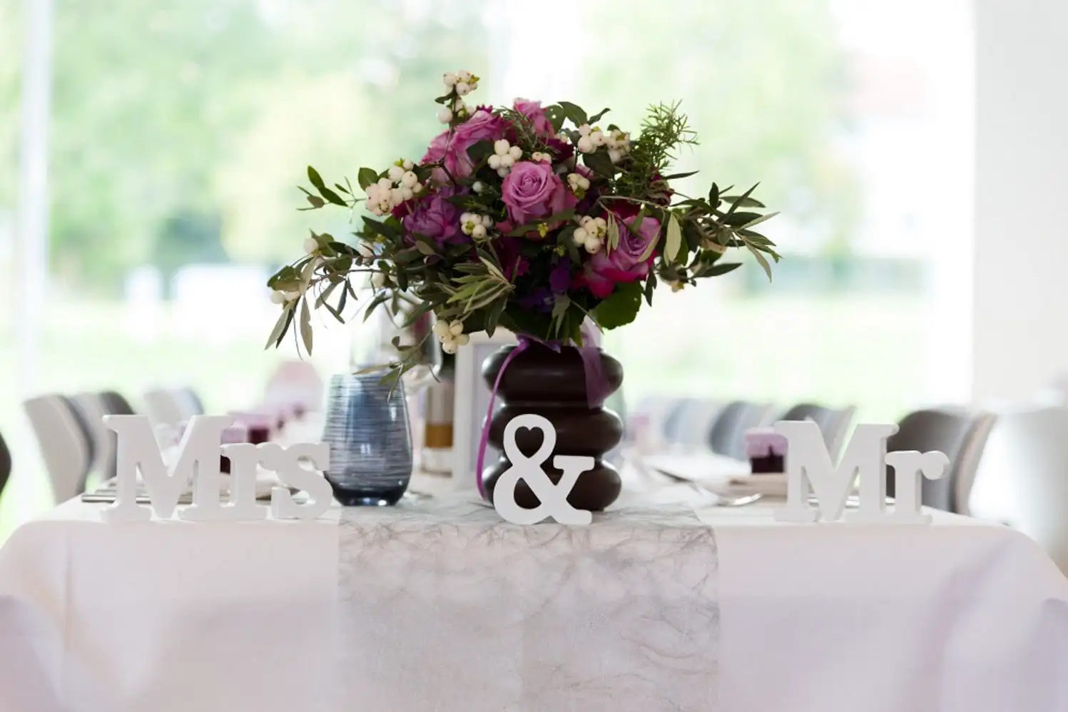 Blumenstrauß mit lila Rosen und weißen Beeren, in einer dunklen Vase auf einem eingedeckten Tisch mit weißen 'Mrs' '&' und 'Mr' Buchstaben darauf.