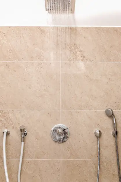 Moderne Dusche mit Wasserstrahl aus quadratischem Duschkopf von oben, die Wand ist beige gefliest und zwei Handbrausen mit Schläuchen sind dort befestigt.