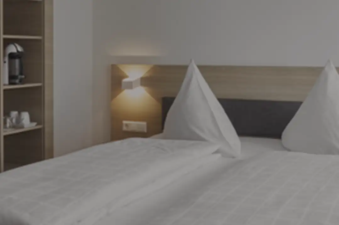 Modernes Hotelzimmer mit Doppelbett, weißen Bettdecken und zu einem Dreieck geformten Kissen, neben einem Holzregal mit Kaffeemaschine und Zubehör.