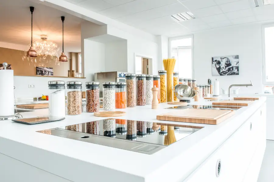 Moderne, helle Küche mit weißen Arbeitsflächen, mehreren Induktionskochfeldern und einer Reihe von transparenten Vorratsgläsern mit verschiedenen Hülsenfrüchten und Nudeln.