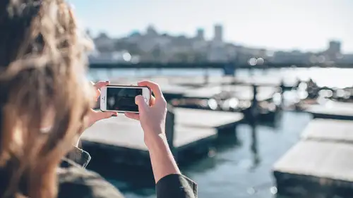 Person hält ein Smartphone und fotografiert einen Hafen in Ulm mit Bootsanlegern im Hintergrund, bei Sonnenlicht.