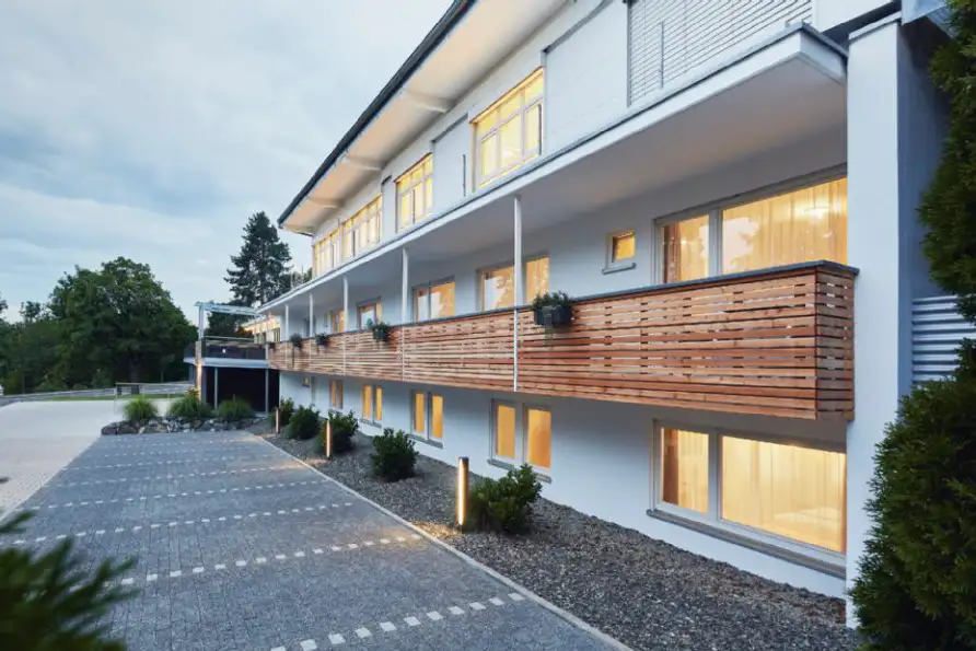 Moderne mehrstöckige Hotelanlage mit beleuchteten Fenstern, hölzernen Balkonen und Parkplätzen, am Abend.