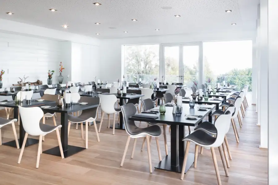 Moderner heller Speisesaal mit weißen Stühlen, darauf graue Polster und gedeckten Tischen vor großen Fenstern mit Blick auf grüne Bäume.