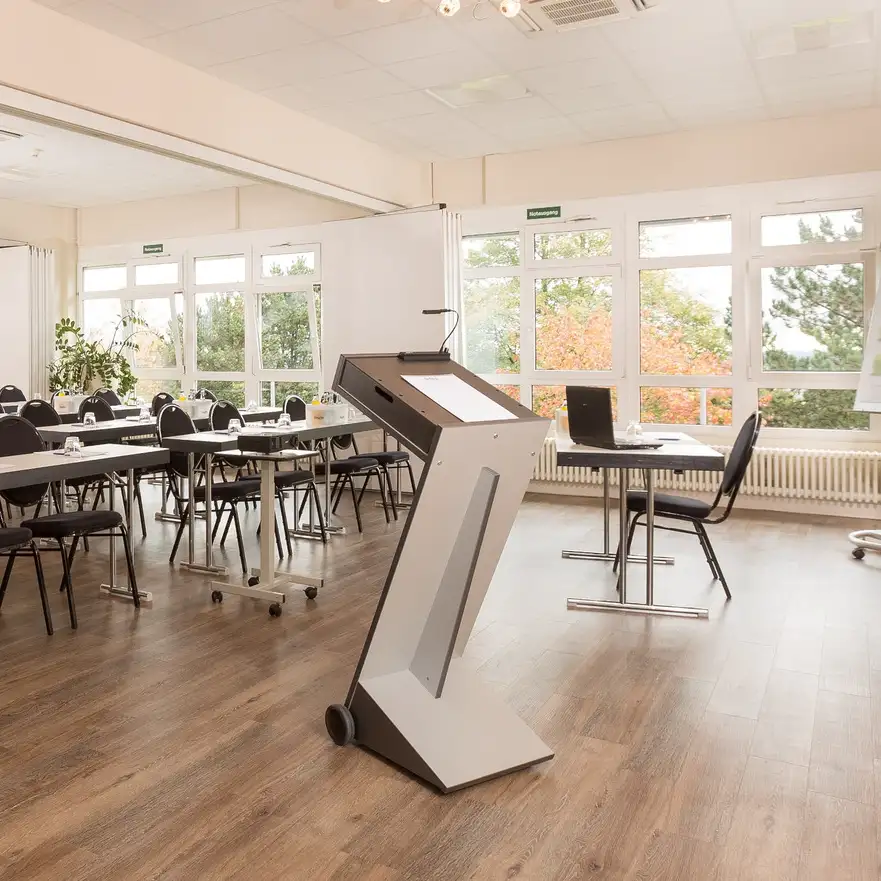 Heller, moderner Konferenzraum mit  Rednerpult mit Mikrofon vor Reihen mit Tischen und Stühlen vor großen Fenstern.