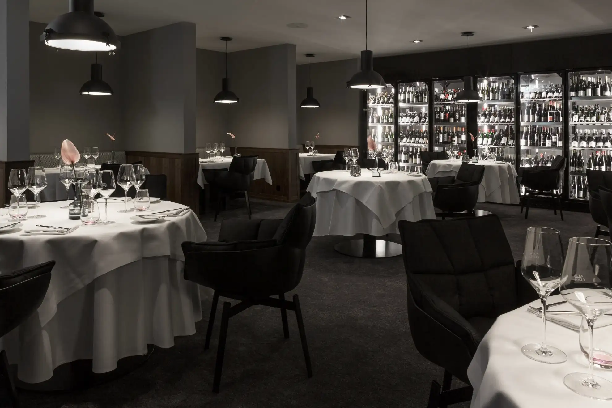 Elegantes Restaurantinterieur mit runden, gedeckten Tischen, weißen Tischdecken, schwarzen Stühlen und beleuchteten Weinregalen an der Wand.