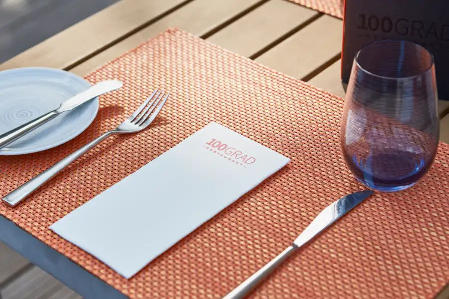 Gedeckter Tisch mit orangenem Tischset, Besteck, blauem Glas und einer Papierserviette vom 100GRAD Restaurant.