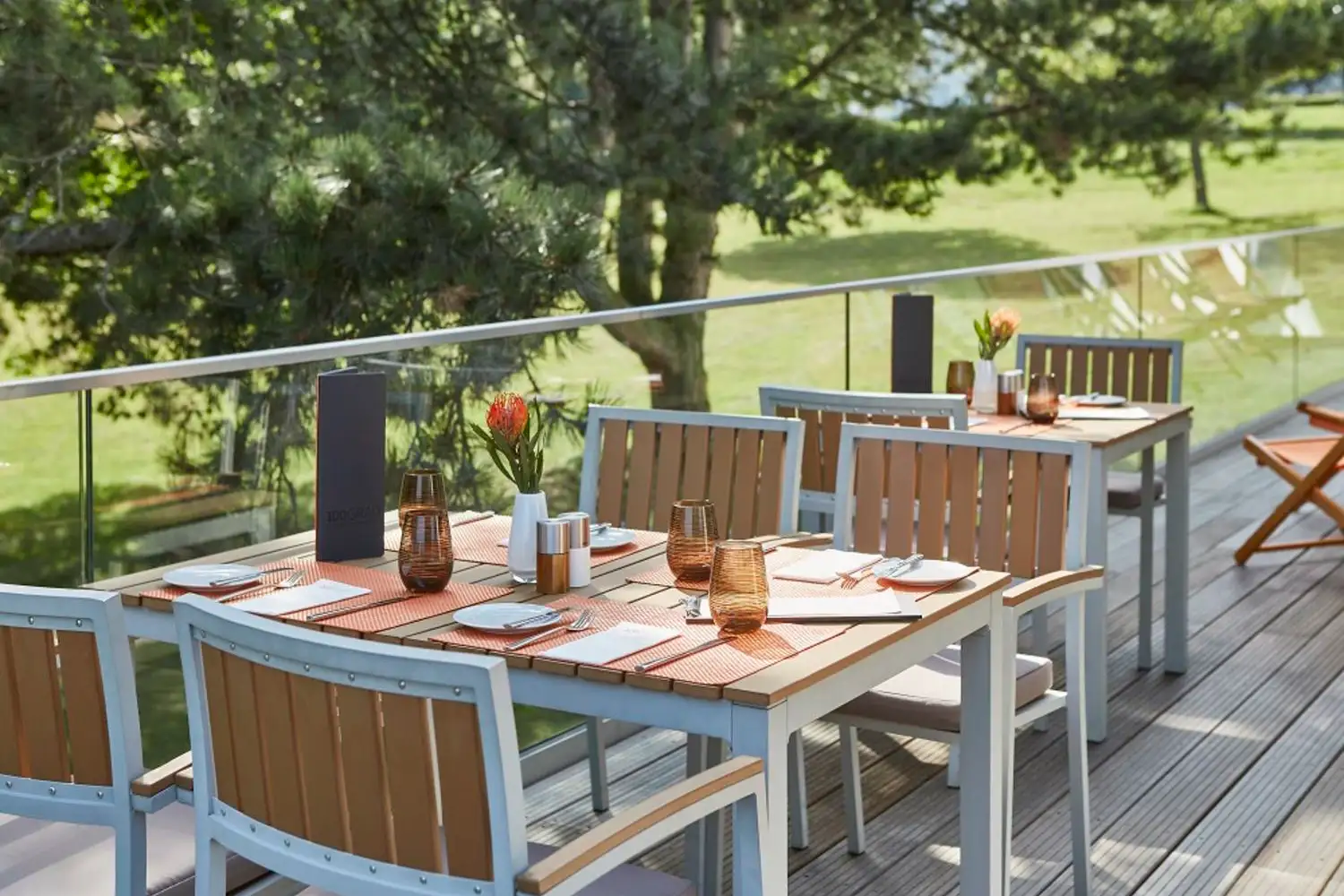 Gedeckte Tische mit orangefarbenen Platzsets und braunen Gläsern auf einer Terrasse mit Blick auf einen grünen Garten.