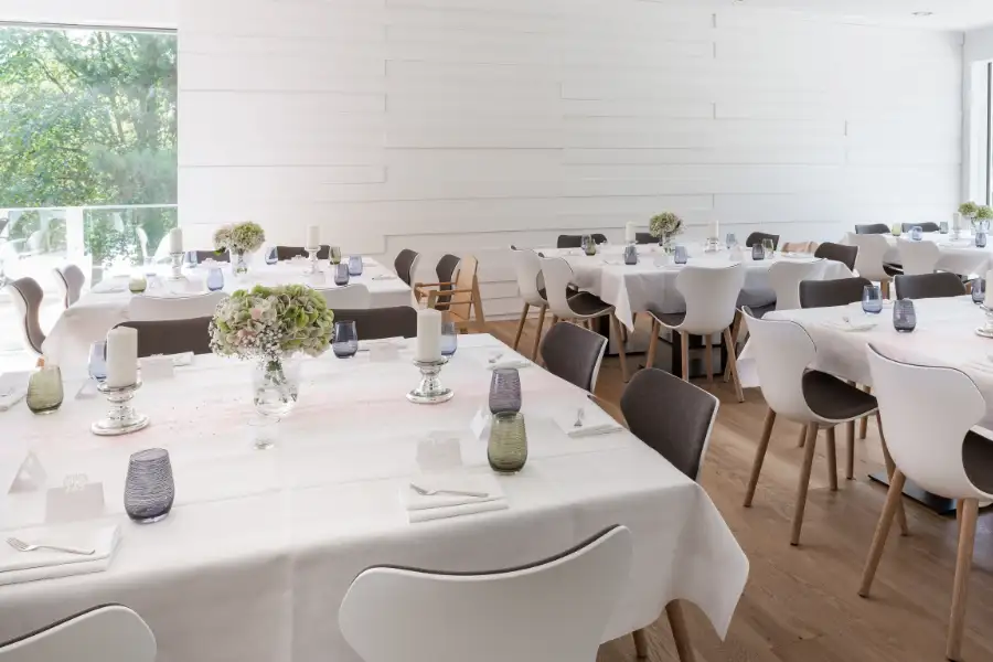 Hell eingerichteter Speisesaal mit weißen Tischtüchern, die  Tische sind elegant eingedeckt mit Kerzenhaltern, Blumenglas und bunten Wassergläsern, die modernen Stühle sind weiß, mit grauen Polstern und es gibt zwei große Fenster.