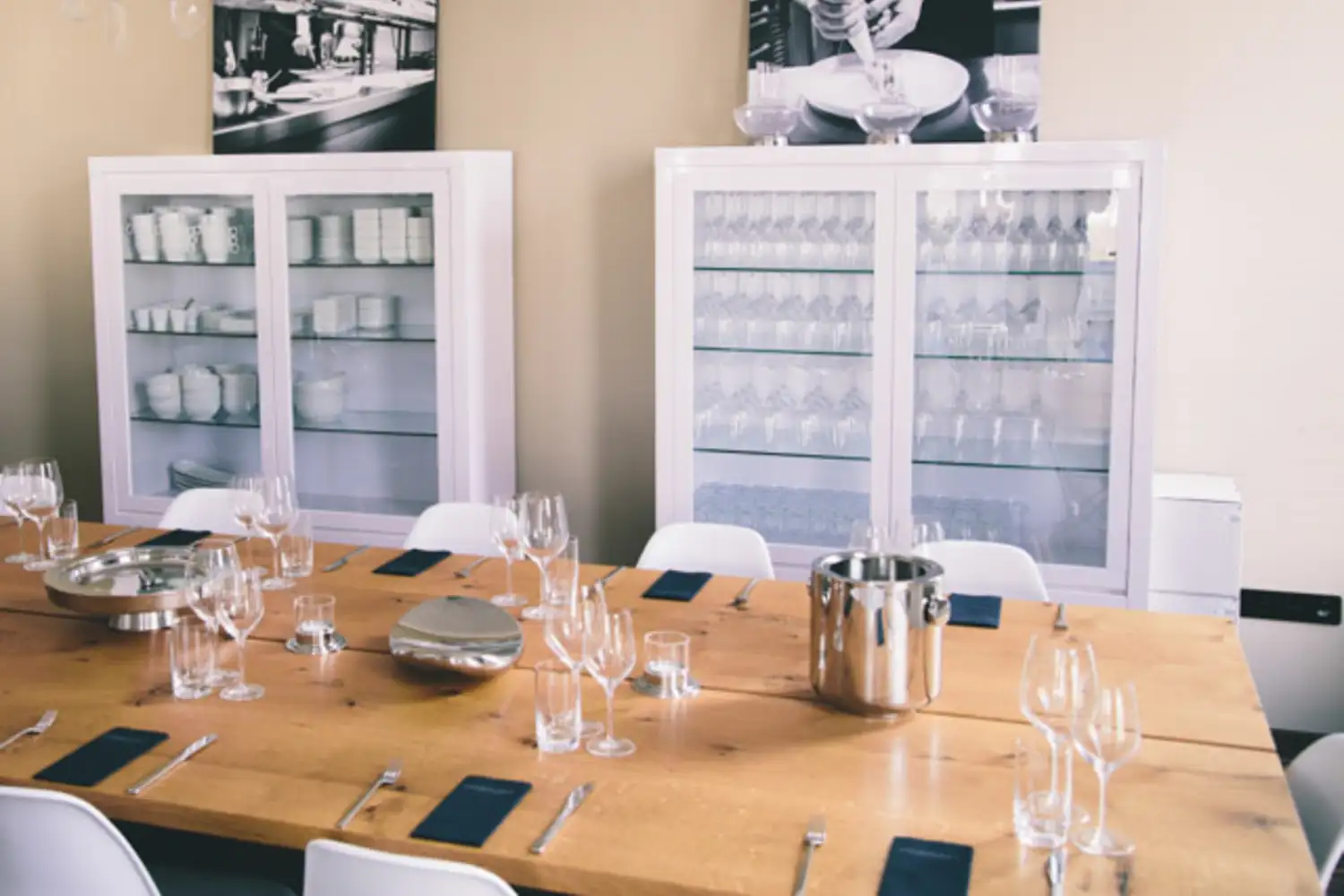 Großer Holztisch mit mehreren Gedecken, Gläsern und silbernen Accessoires in einem hellen Raum mit weißen Küchenvitrinen im Hintergrund, die mit Gläsern und Geschirr befüllt sind.