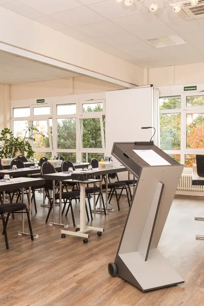 Heller, moderner Konferenzraum mit  Rednerpult mit Mikrofon vor Reihen mit Tischen und Stühlen vor großen Fenstern.