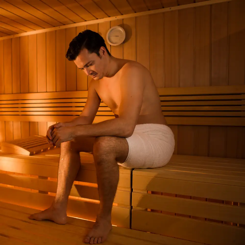 Ein Mann sitzt in einer Sauna auf einer Holzbank und trägt ein weißes Handtuch um die Taille, im Hintergrund hängt eine Uhr.