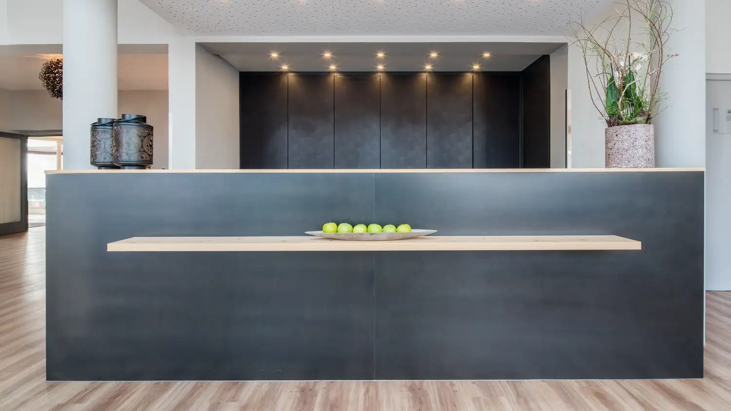 Moderne Rezeption mit schwarzer Front, Holzablage, grünen Äpfeln in einer Schale, zwei Gefäßen links und einem Blumentopf mit Zweigen rechts.