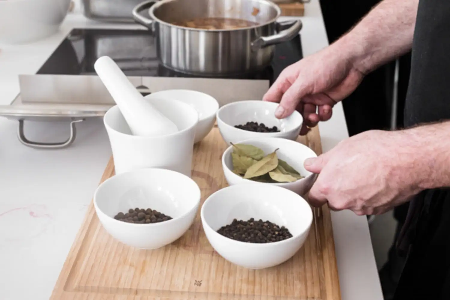 Zwei Hände eines Kochs halten eine weiße Schüssel mit Lorbeerblättern neben anderen Schüsseln mit Gewürzen und einem Mörser auf einem Holzbrett in einer Küche.