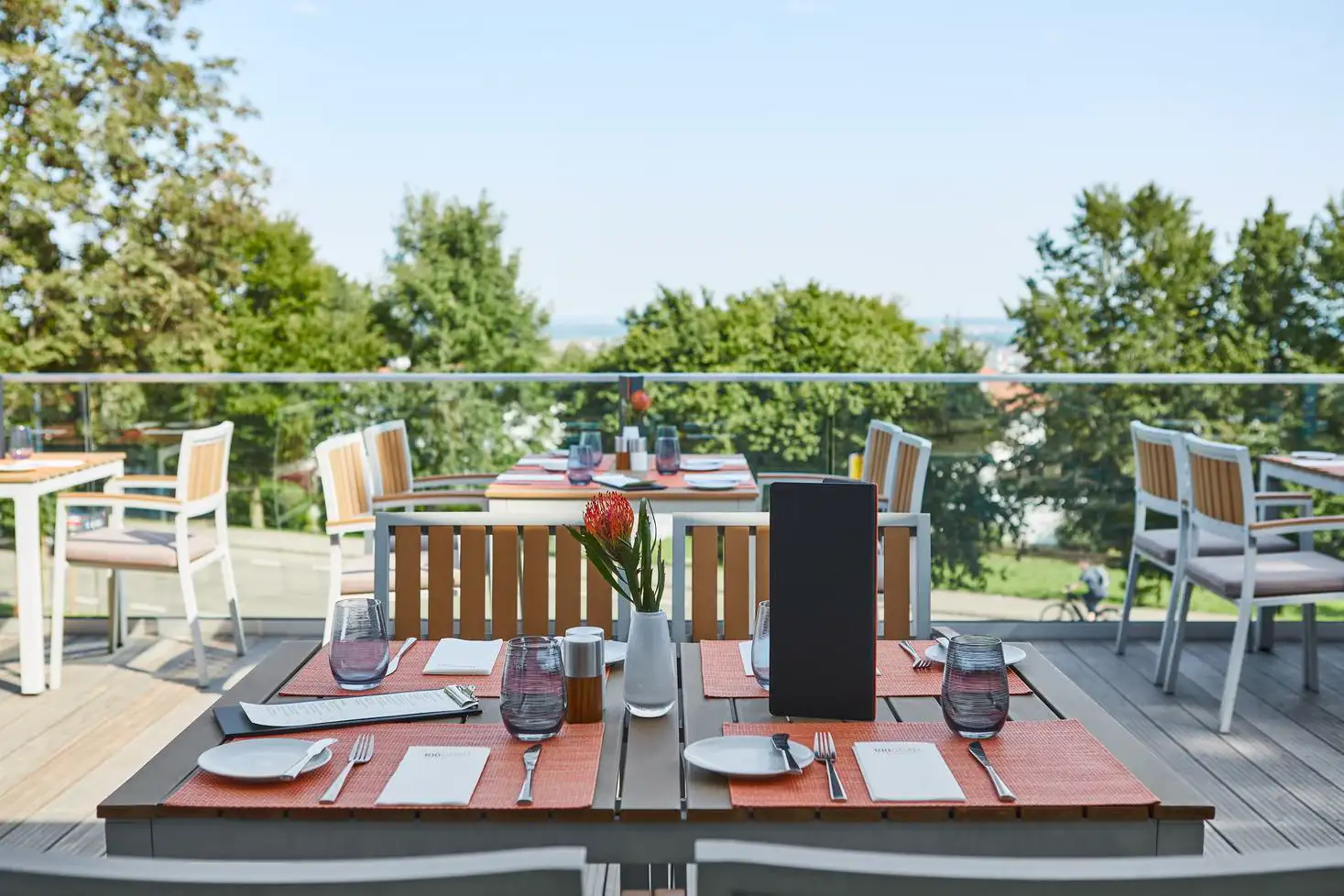 Sonnbelichtete Außenterrasse eines Restaurants mit eingedeckten Tischen, Stühlen und Blick auf grüne Bäume.